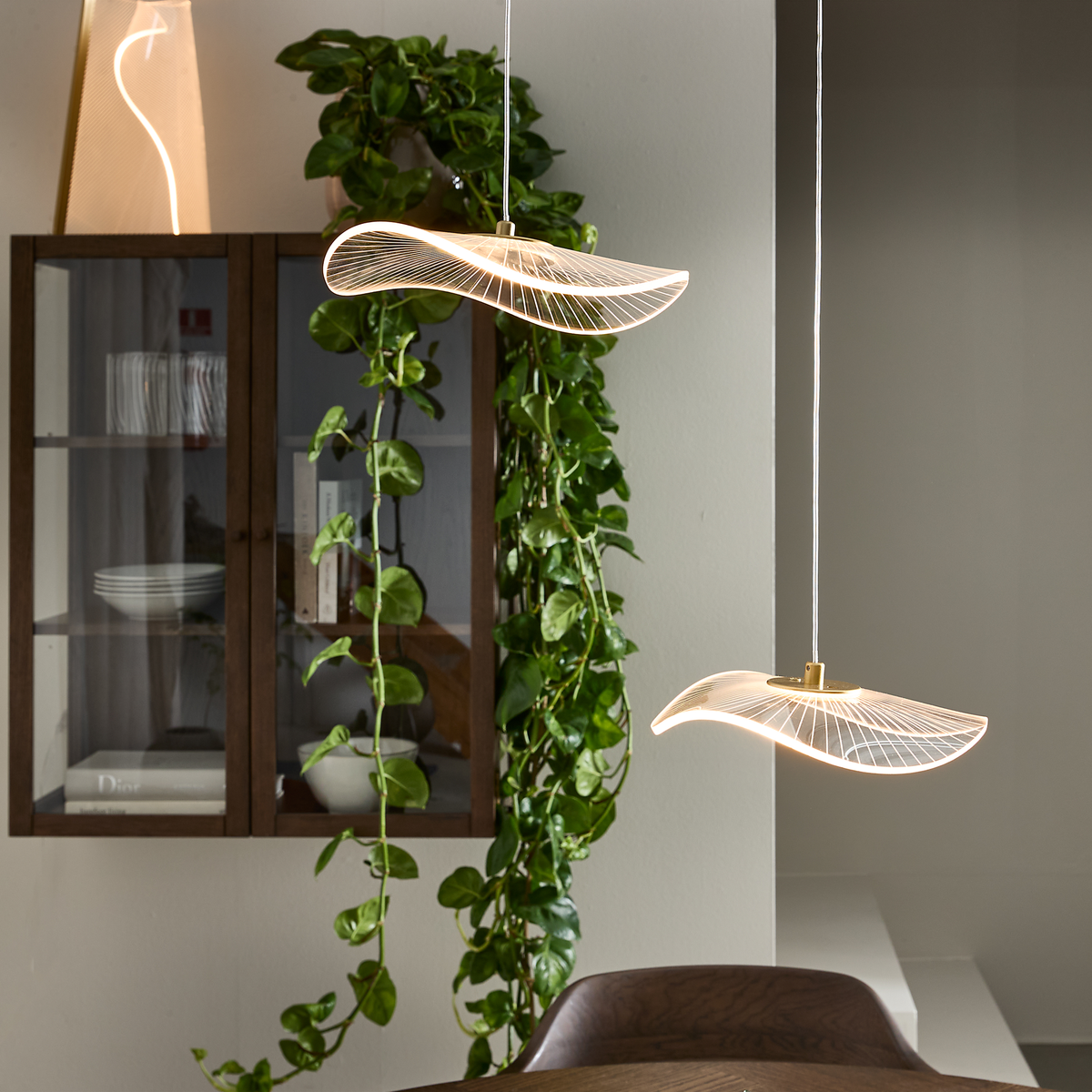 To skulpturelle bølgeformede pendler over et spisebord, varm belysning, vitrineskab i træ og nedhængende grøn plante.