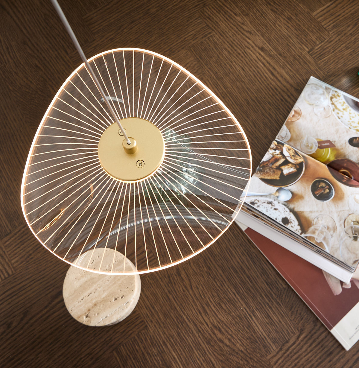Moderne gulvlampe med transparent skærm og radiale linjer, messingdetalje og rund stenfod på mørkt trægulv ved siden af et åbent magasin.