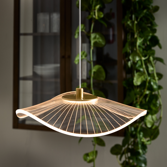 Bølgeformet pendellampe med varm LED, transparent skærm og guldfarvet top, hænger foran vitrineskab og grønne klatreplanter.