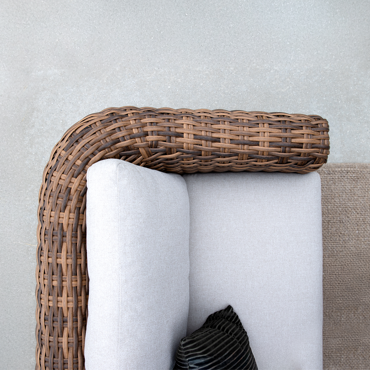 Nærbillede af hjørnet på en rattan-sofa med lysegrå puder og en lille sort pude, udendørs i dagslys.