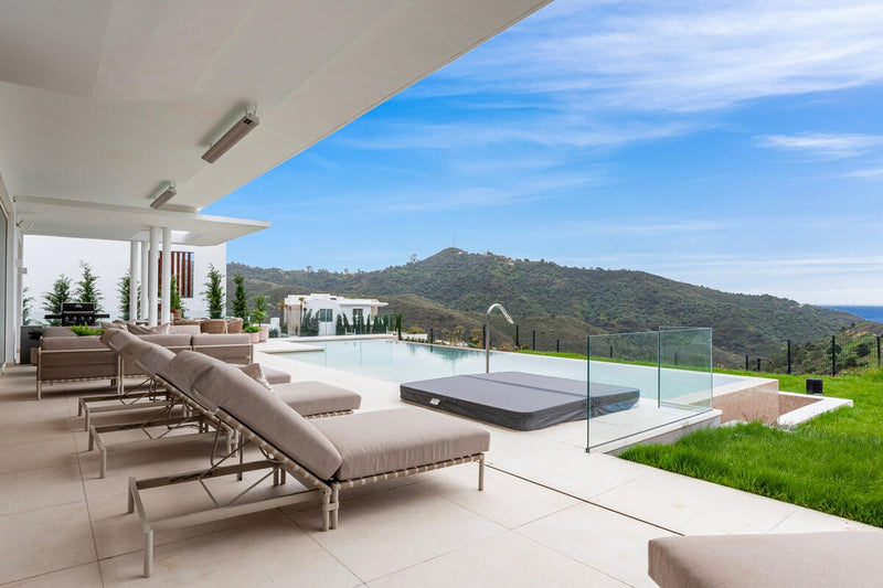 Moderne terrasse med solsenge, infinitypool og udsigt til grønne bjerge under blå himmel.