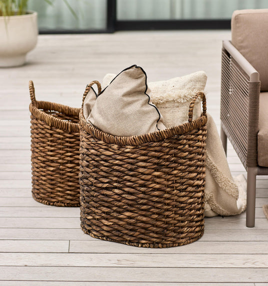 To flettede opbevaringskurve på en lys træterrasse, fyldt med beige puder og et tæppe ved siden af en udendørs sofa.