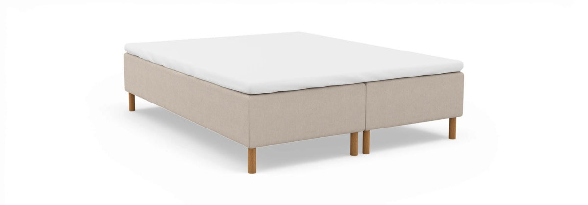 Beige kontinentalsengbund med hvid topmadras og runde træben, frilagt på hvid baggrund.