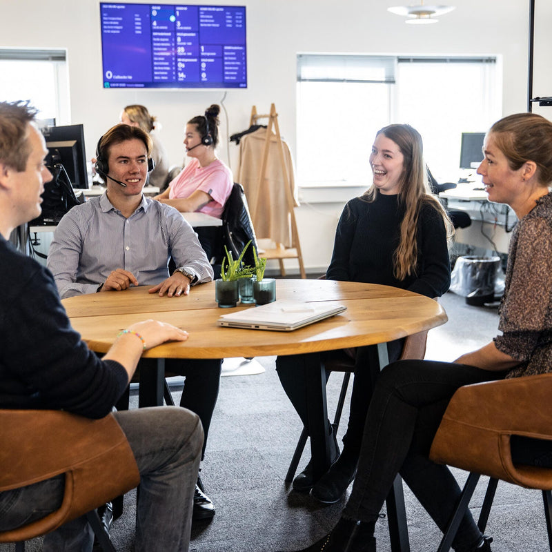 Gruppe­møde ved rundt træbord på moderne kontor med bærbar computer og små grønne planter; kolleger og arbejdsstationer i baggrunden samt dashboards på vægskærm.