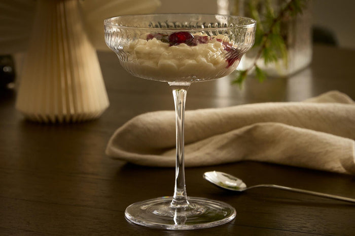 Dessert serveret i et riflede coupe-glas på et mørkt træbord med linnedserviet og ske ved siden af; sløret vase med grønne grene og en papirengel i baggrunden i varm belysning.