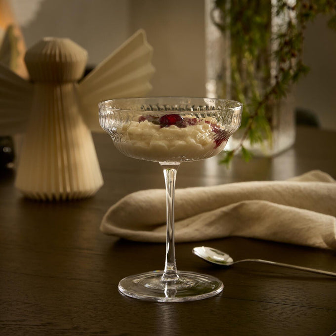 Dessert serveret i et riflede coupe-glas på et mørkt træbord med linnedserviet og ske ved siden af; sløret vase med grønne grene og en papirengel i baggrunden i varm belysning.