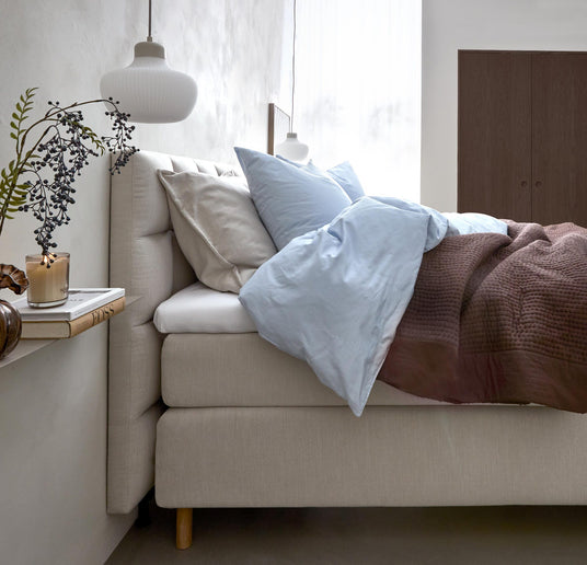 Lyst soveværelse med beige polstret seng, lyseblåt sengesæt og brun strikket plaid, pendellamper over samt hylde med vase, bog og tændt lys.