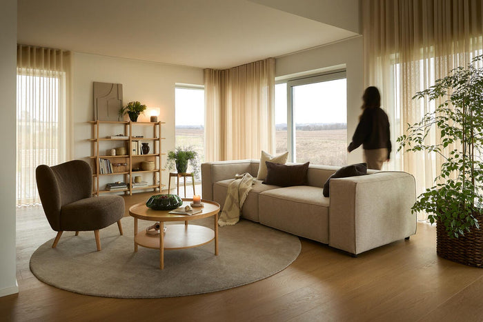 Lyst skandinavisk stue med beige hjørnesofa, brun lænestol og rundt tæppe; store vinduer med lette gardiner, træreol og træbord med grøn skål og tændt lys; en person i bevægelse ved vinduet.