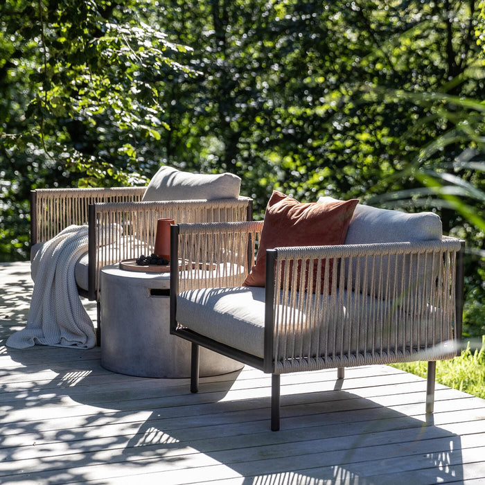 To udendørs lænestole med snore-detaljer og lyse hynder på en træterrasse ved siden af et rundt sidebord i beton, med plaid og rustfarvet pude, omgivet af solbelyst grønt.