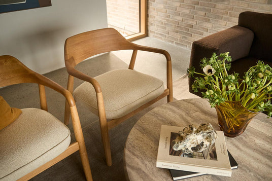 Stenbord med bogen 'barefoot living' og en sten som papirvægt ved siden af en ravfarvet vase med hvidgrønne blomster; to træloungestole med lyse hynder og en brun sofa i en lys stue.