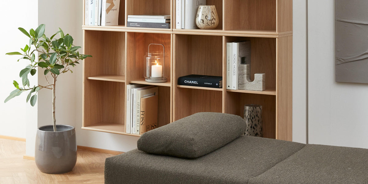 Lyst stue med reol i eg med bøger som PRADA og CHANEL, vaser og en tænd lyslygte, ved siden af en grå daybed, gulvlampe og potteplante.
