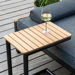 Lille sidebord i træ med sort metalstel ved siden af en grå loungesofa på træterrasse; et glas hvidvin på bordet og en åben bog samt puder på sofaen.