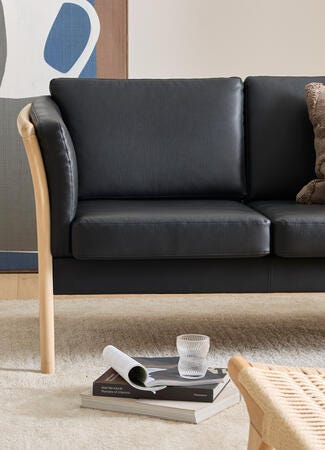 Sort lædersofa med lys treramme og en beige, tekstureret pude; foran ligger en stak coffeetable-bøger – bl.a. ”Axel Vervoordt: Portraits of Interiors” – med et riflede glas ovenpå; vævet loungestol og neutral tæppe i en lys stue.