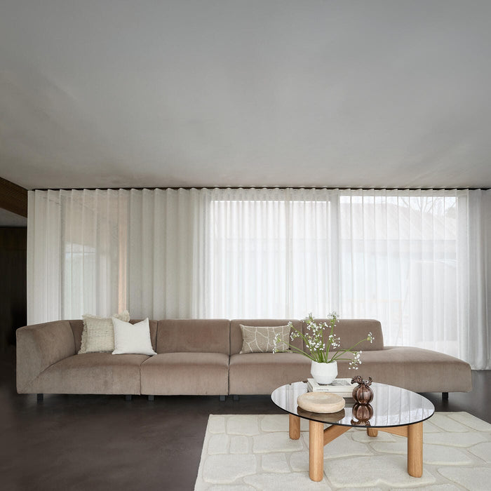 Lys stue med beige modulsofa, lette hvide gardiner, rundt glasbord med træben og en vase med hvide blomster på lys tæppe.