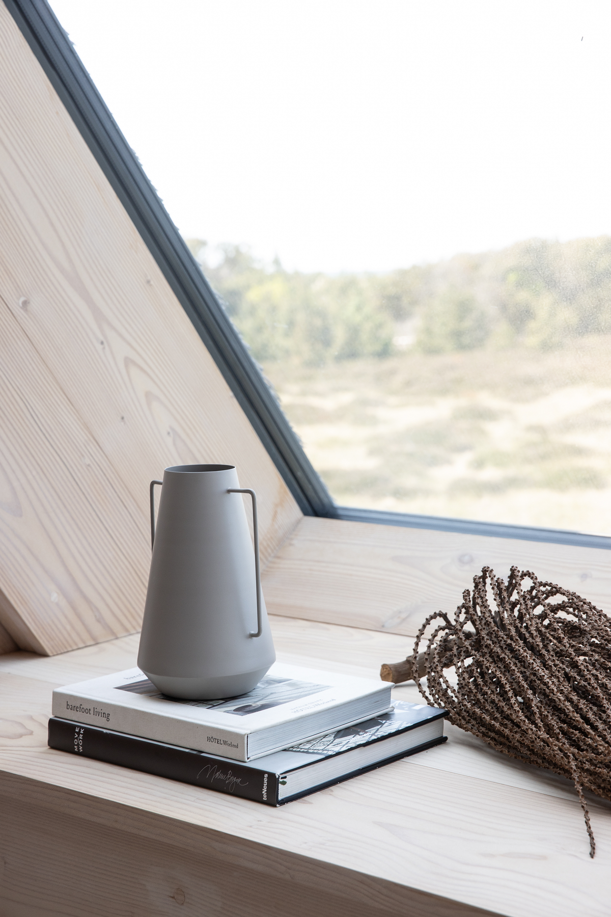 Grå kande på tre bøger: “barefoot living”, “HOTEL WEEKEND” og “Monochrome”, på en lys trævindueskarm med udsigt over naturen; ved siden ligger en tørret gren.