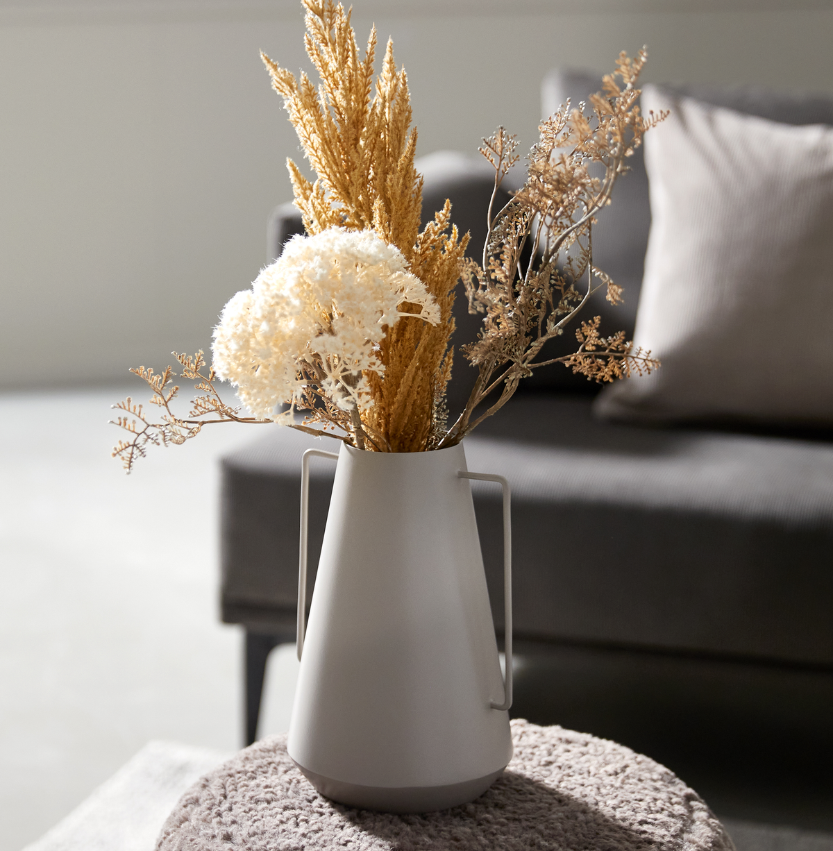 Lys grå vase med tørrede blomster på en puf foran en sofa i blødt dagslys.