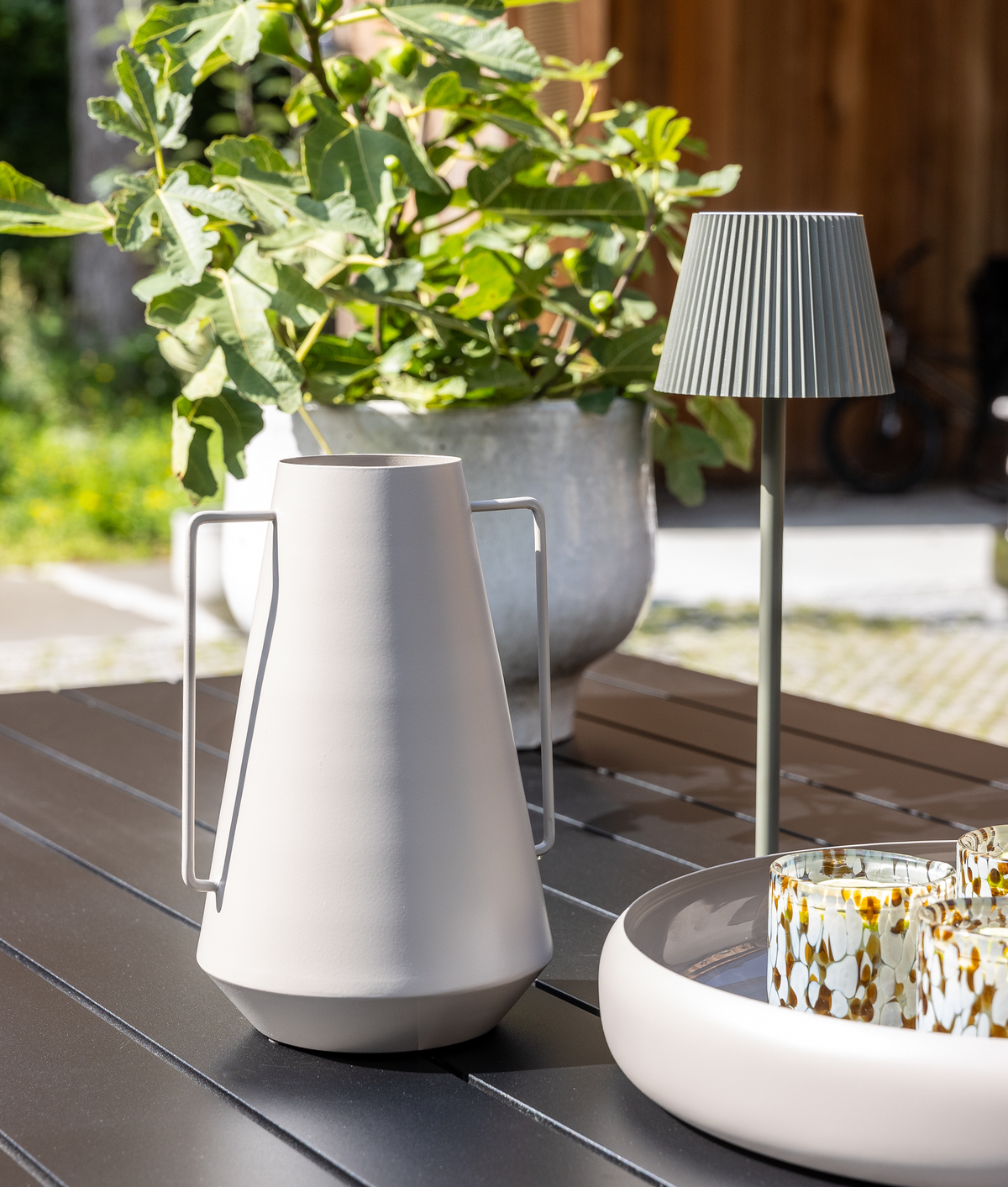 Moderne beige kande/vase og bærbar bordlampe på et udendørs bord; bakke med mønstrede glas og en potteplante i baggrunden.