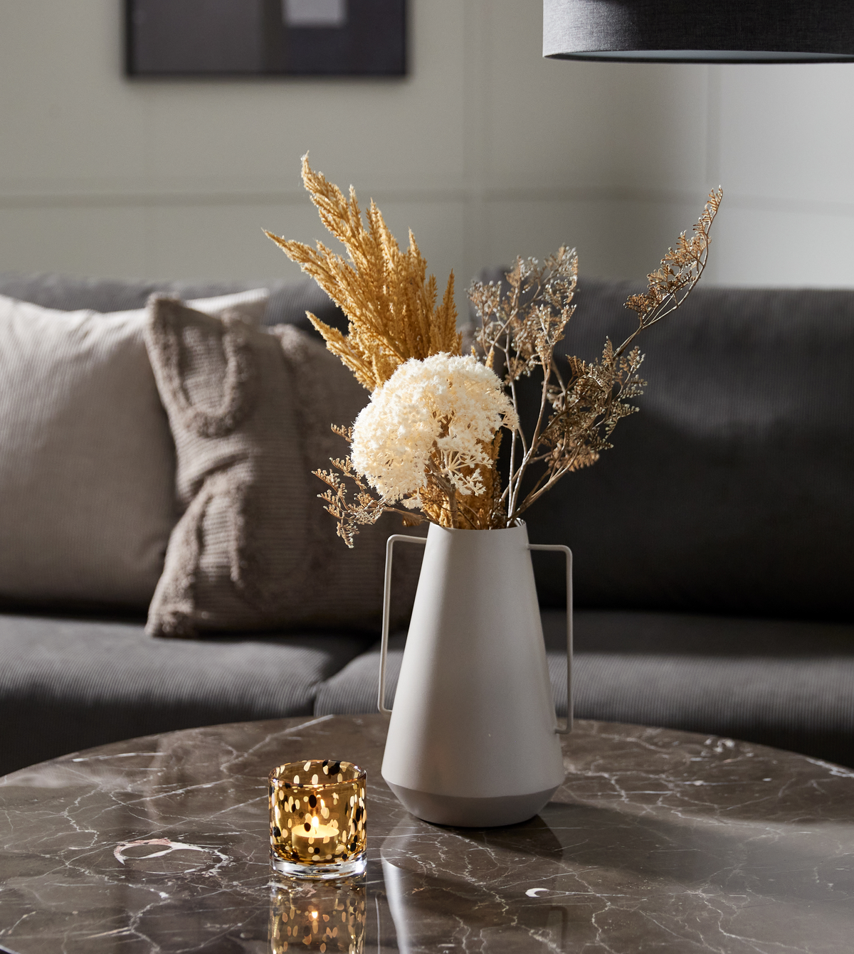 Grå sofa med puder bag et marmorbord med tændt fyrfadsstage og grå vase med tørrede blomster.