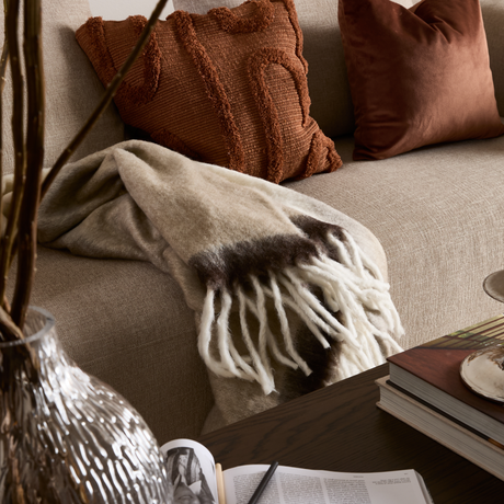 Beige sofa med rustfarvede puder, uldtæppe med frynser i beige og brunt, glasvase med grene og bøger på et mørkt sofabord – hyggeligt stuehjørne.