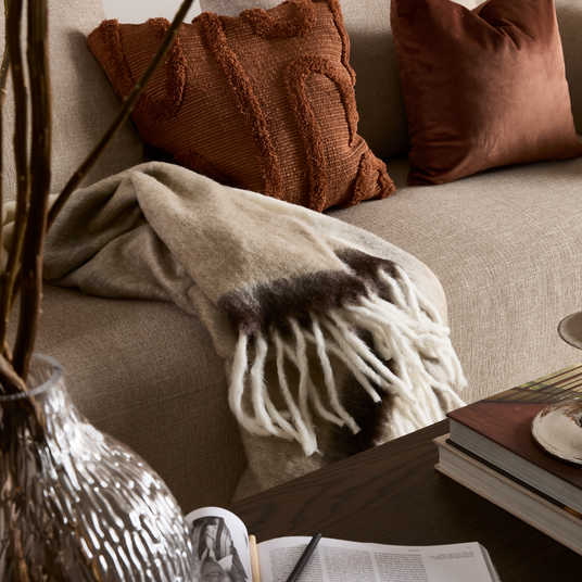 Beige sofa med rustfarvede puder, uldtæppe med frynser i beige og brunt, glasvase med grene og bøger på et mørkt sofabord – hyggeligt stuehjørne.