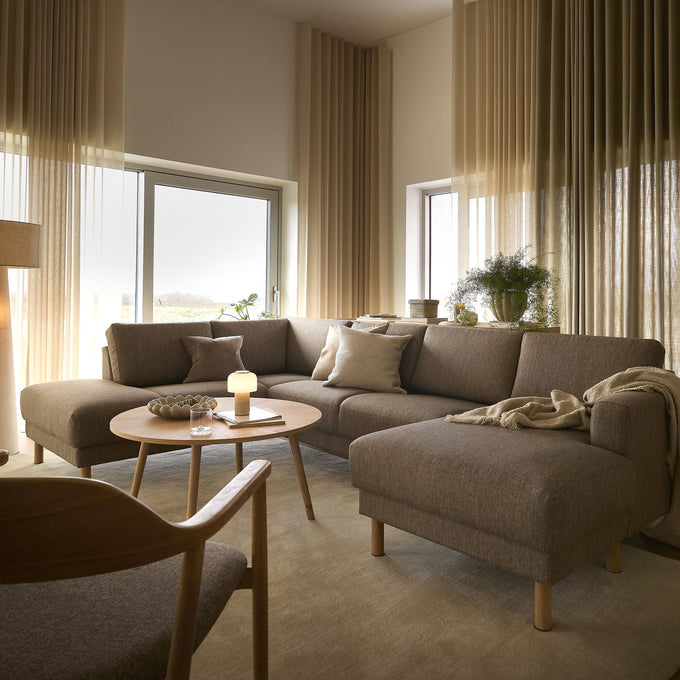 Hyggelig skandinavisk stue med beige gardiner, brun hjørnesofa med chaiselong, rundt sofabord i træ med tændt lampe, plaid og puder, tæppe og gulvlampe ved store vinduer i dagslys.