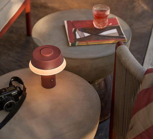 Bærbar bordlampe i vinrød, tændt på et rundt betonbord ved siden af et kamera; på bordet bagved står et glas med rød drik og en bog i en hyggelig stue.