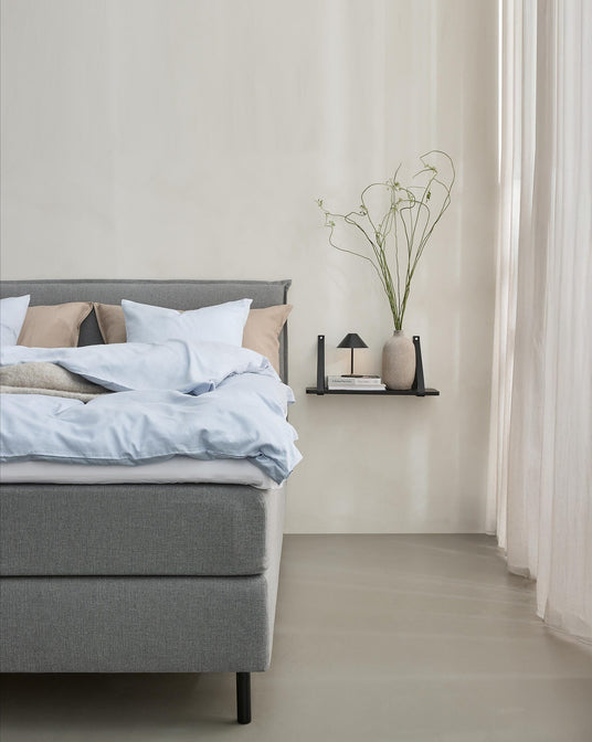 Minimalistisk soveværelse med grå seng, lyseblåt sengetøj og beige puder; ved siden af er en sort væghylde med en lille lampe, bøger og en vase med lange kviste ved lyse gardiner.