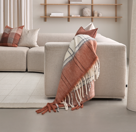 Beige sofa med et rust- og hvidt ternet uldtæppe med frynser lagt over armlænet; puder, lyse gardiner og træhylder i baggrunden.