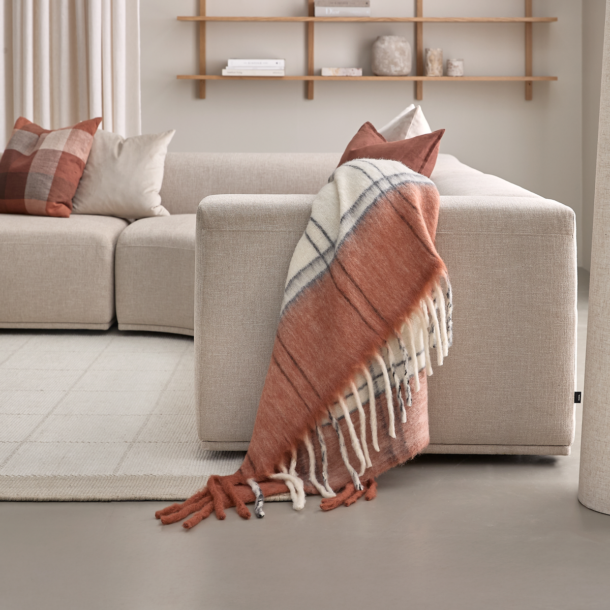 Beige sofa med rust- og cremefarvet ternet plaid med frynser over armlænet, matchende puder og åbne hylder i en lys stue.