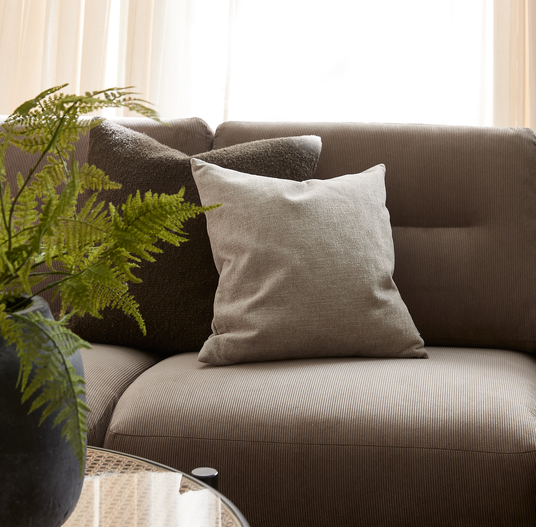 Nærbillede af en beige-brun sofa med to pyntepuder, én lys og én mørk tekstureret, en grøn bregne i sort vase og bløde lyse gardiner i dagslys.