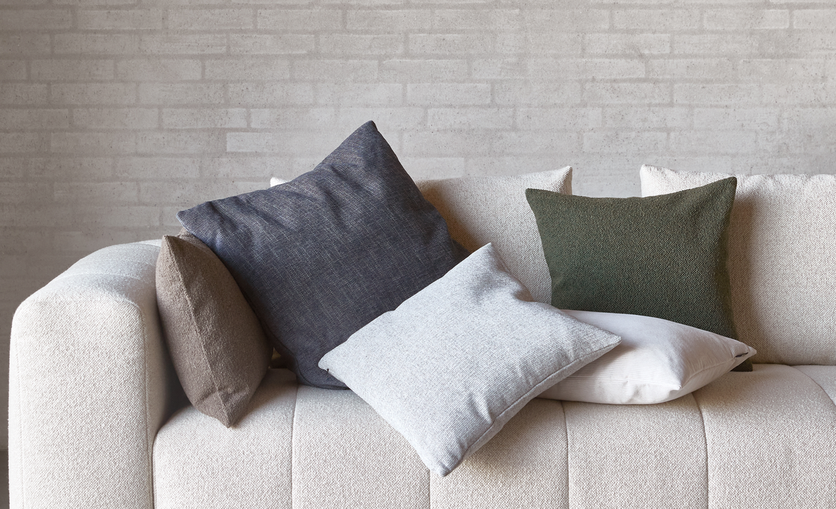 Beige sofa med dekorative puder i grå, grøn, hvid og brun foran en lys murstensvæg.