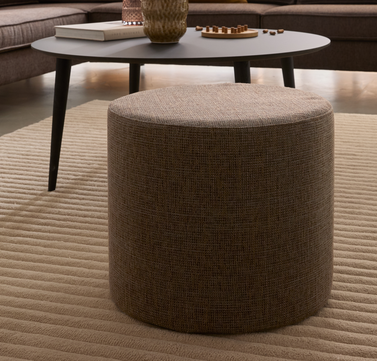 Beige cylinderformet puf på en rillet beige tæppe foran et mørkegråt sofabord med bog, vase og lille spillebrikbakke.
