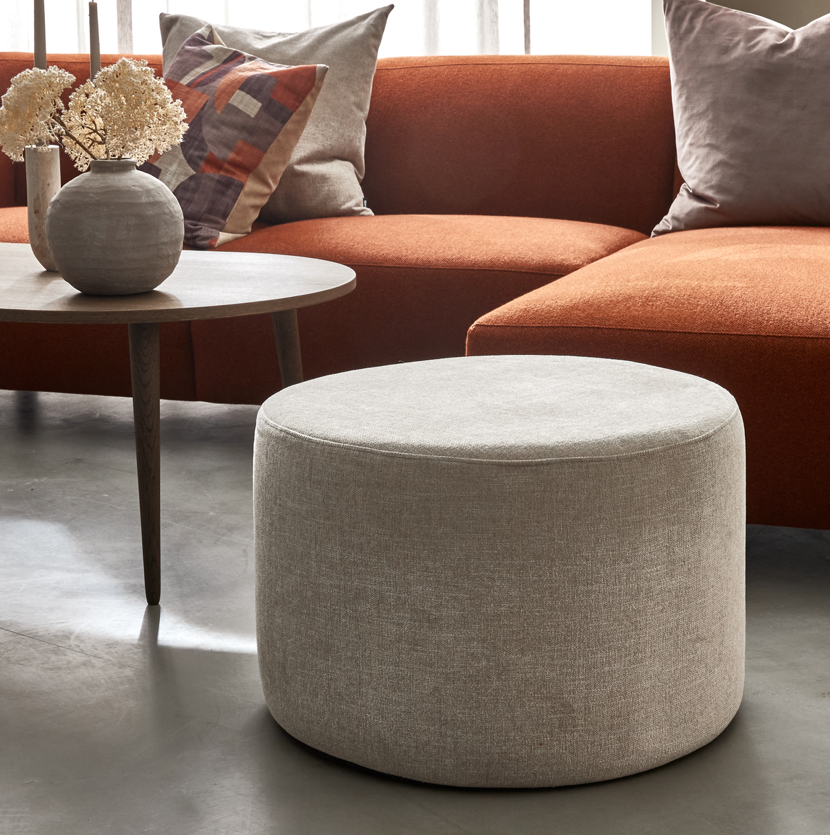 Beige, rund puf i moderne stue med rustfarvet sofa, træ-sofabord med vase og lys samt neutrale puder.