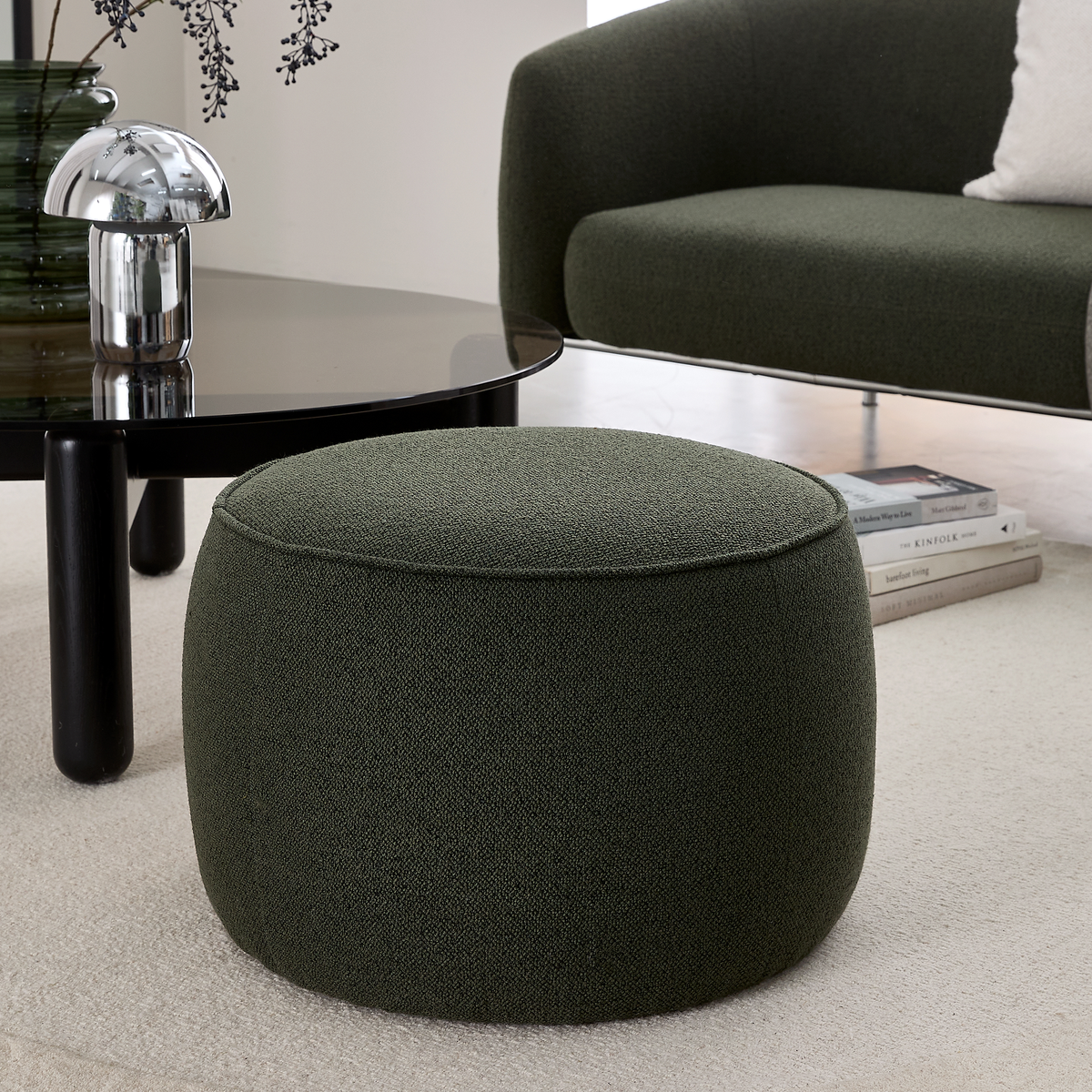 Mørkegrøn rund puf i en moderne stue ved siden af et sort glas sofabord med krom bordlampe og sofa på lys måtte.