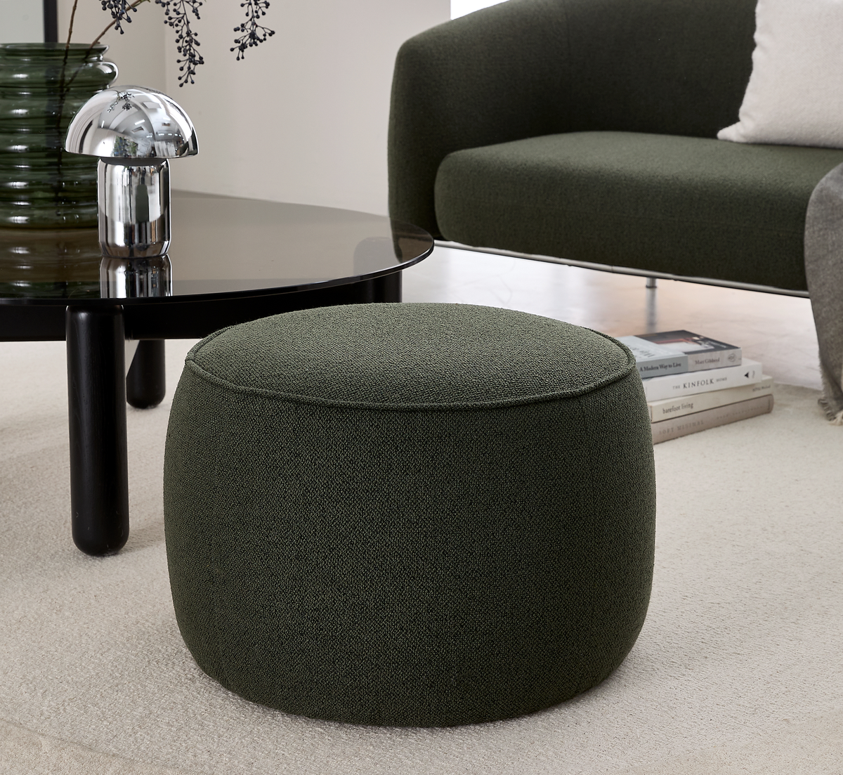 Mørkegrøn rund puf ved et sort sofabord med krom svampelampe; grøn sofa og lyst tæppe i en moderne stue.