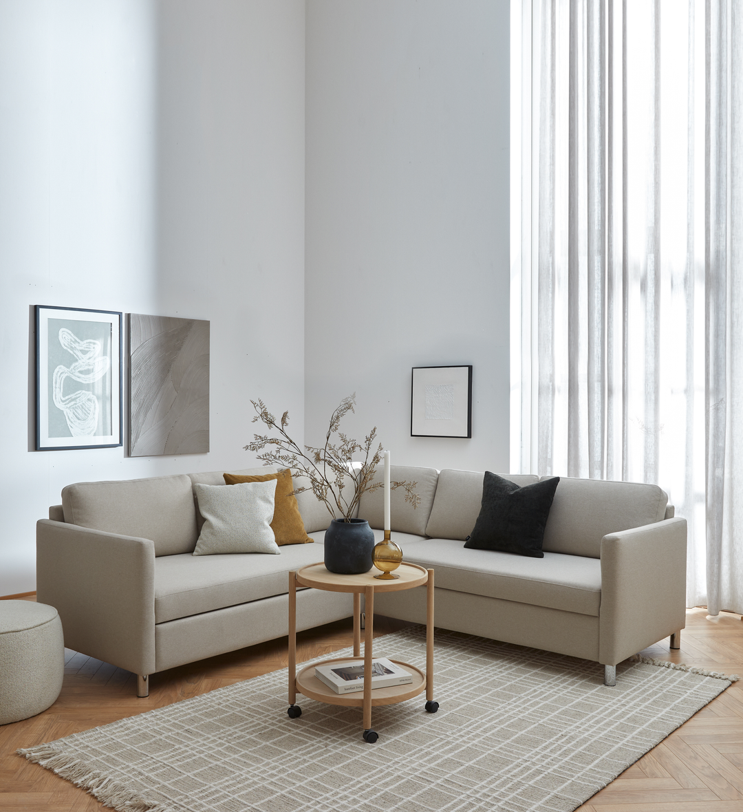Lyst stue i skandinavisk stil med beige hjørnesofa, rundt rullebord med vase, lys og tørrede grene, ternet tæppe, vægkunst og lette gardiner ved stort vindue.