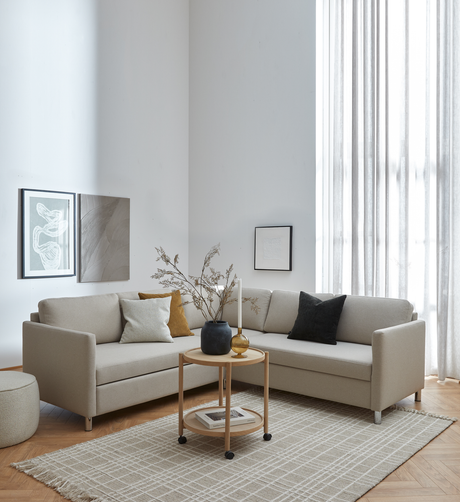 Lyst stue i skandinavisk stil med beige hjørnesofa, rundt rullebord med vase, lys og tørrede grene, ternet tæppe, vægkunst og lette gardiner ved stort vindue.
