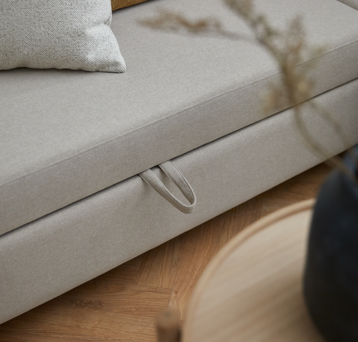 Nærbillede af beige sofa med træksstrop på underdelen, pude ovenpå og rundt sofabord i træ med mørk vase sløret i forgrunden på sildebensparket.