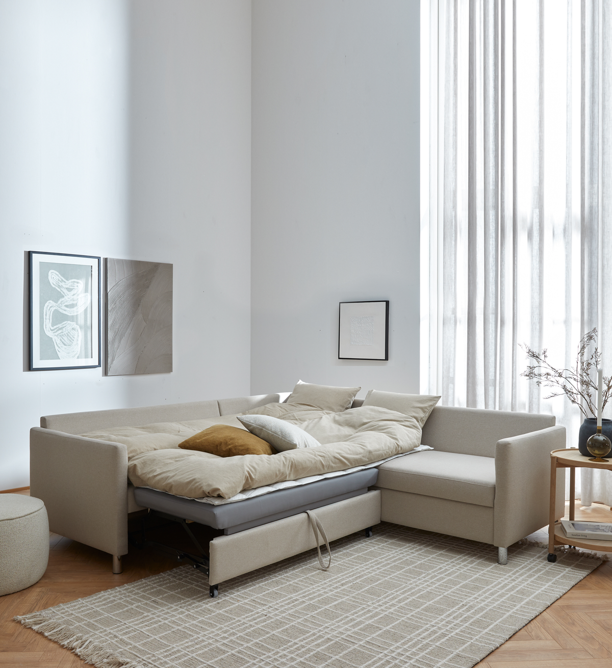 Beige L‑formet sovesofa trukket ud som seng i en lys skandinavisk stue med stor vinduesvæg, lette gardiner, neutral tæppe og sidebord med vase.