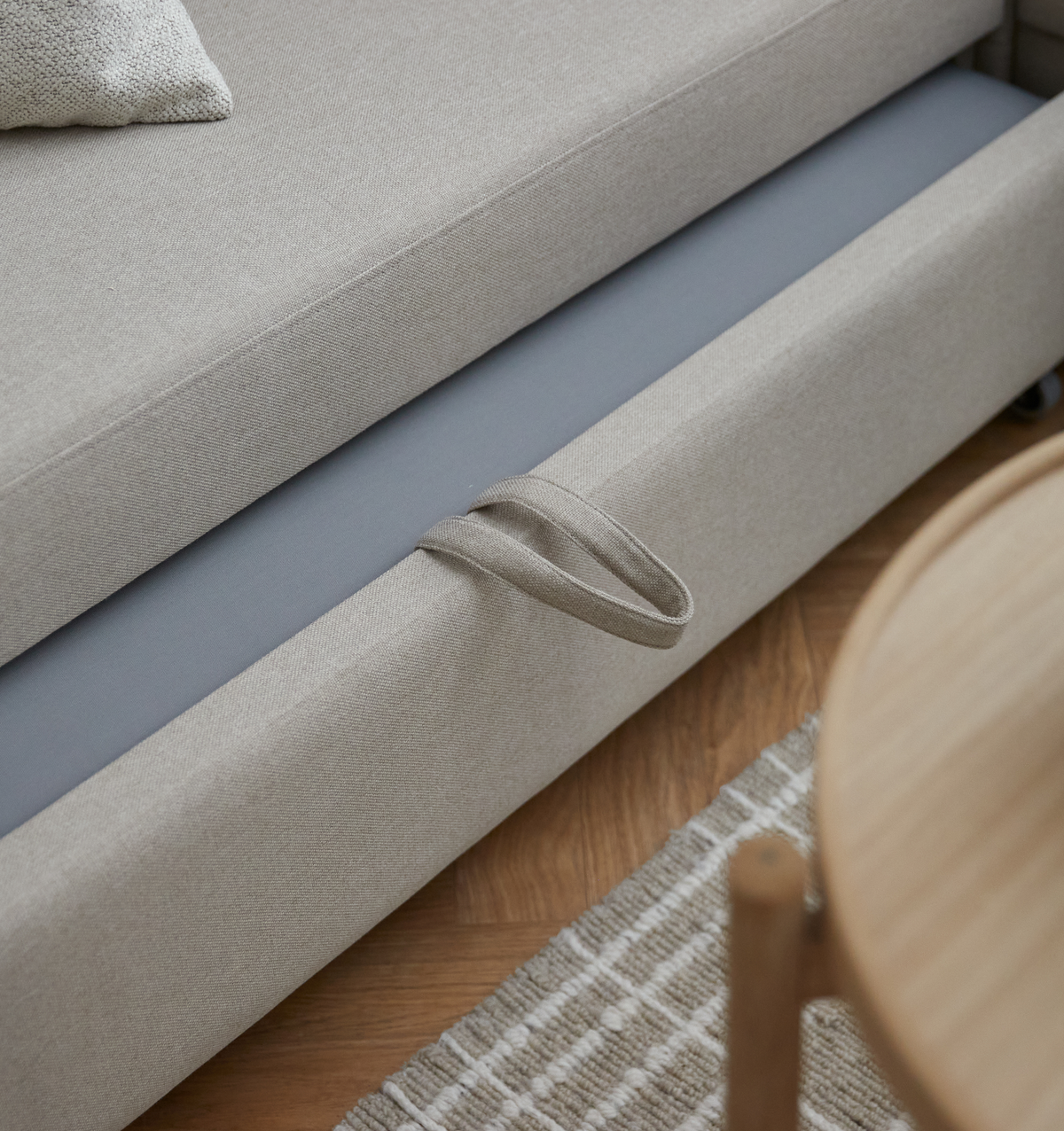 Nærbillede af en beige stofsofa med trækstrop på bunden og grå panel, ved siden af trægulv, en stribet teksturtæppe og kanten af et rundt træbord.