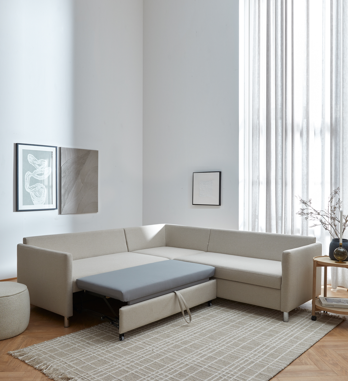 Beige L-formet hjørnesofa med udtræksseng trukket frem i en lys, minimalistisk stue med lette gardiner, neutral ternet tæppe, vægkunst og rundt sidebord med vase.