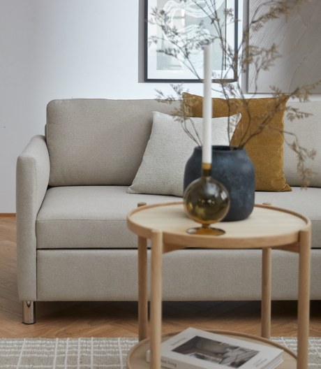 Beige sofa med neutrale og sennepsgule puder, rundt træbord med lys, mørk vase med tørrede grene og indrammet kunst i en lys stue.