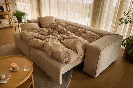 Beige sovesofa slået ud med sandfarvet sengetøj i en lys stue med lette gardiner, rundt træbord med tændt lys og grønne planter.