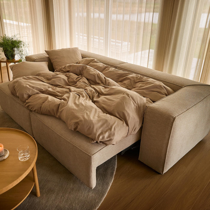 Beige sovesofa slået ud med sandfarvet sengetøj i en lys stue med lette gardiner, rundt træbord med tændt lys og grønne planter.