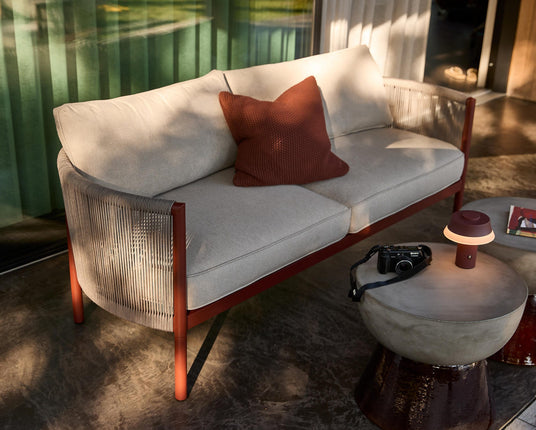 Solbelyst udendørs sofa med rustfarvet stel og beige hynder, en rustrød pude samt to runde sideborde med en bærbar bordlampe og et kamera på en betonterrasse.
