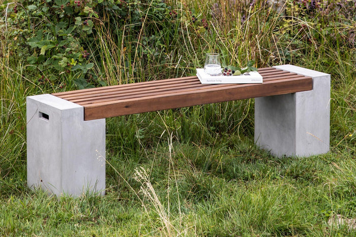 Udendørs bænk med betonben og trælameller, med et glas vand og bogen ”barefoot living” ovenpå i græsset.