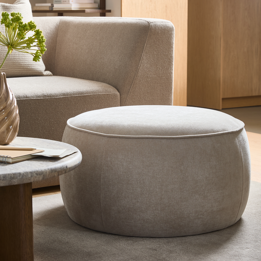 Rund beige stofpuf i en moderne skandinavisk stue ved siden af en buet sofa og et stenagtigt sofabord.