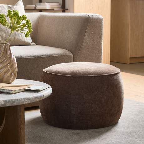 Brun rund puf foran beige sofa, marmorbord med bølget vase og grønne stilke i en lys stue.