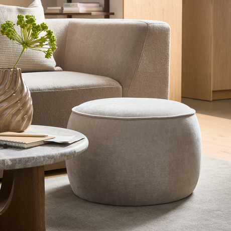 Beige, rund puf i stof foran en buet sofa, med stuebord i sten, bølget vase med grønne stilke og bøger i en lys stue.