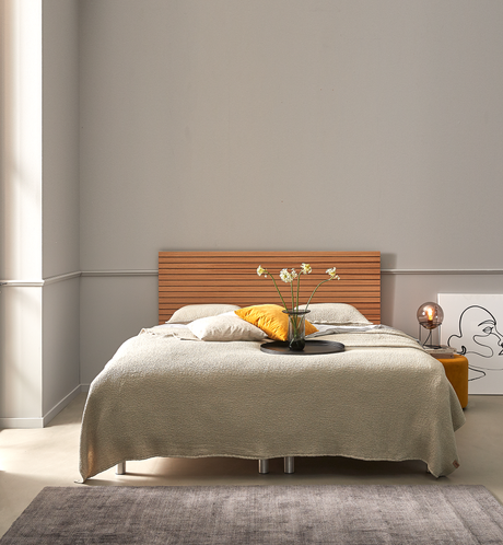 Soveværelse med lav seng og trægavl, beige sengetæppe, hvide og gule puder, vase med blomster på sort bakke, bordlampe og lineart-plakat mod lysgrå væg.